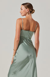 A woman is wearing a light green satin slip dress with thin spaghetti straps. The dress features a fitted bodice with delicate lace or trim detail along the top edge. The fabric appears smooth and shiny, with a flowing, elegant silhouette. She is accessorized with gold hoop earrings and gold bangles on her wrist, and her hair is styled in a neat, low bun.